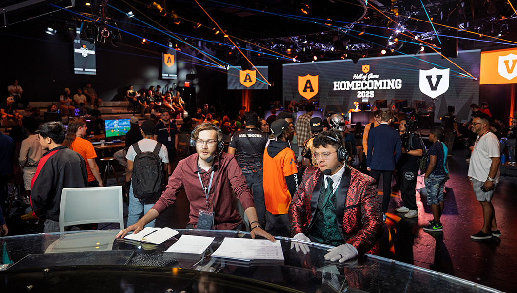 The event room on the day: casters at the desk in front of a crowd, with A and V shield banners on the walls and the Homecoming 2025 walk-in screen on the main display