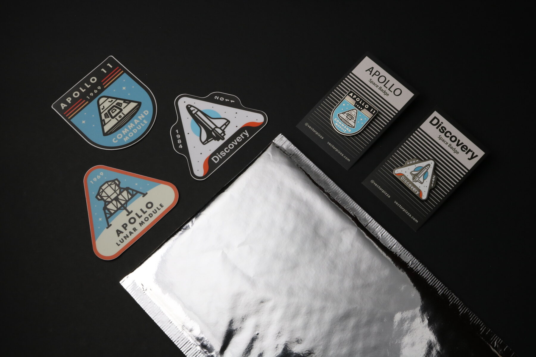 Flat lay of Apollo 11, Discovery and Lunar Module stickers, the Apollo and Discovery pins on backing cards, and a silver mylar mailer