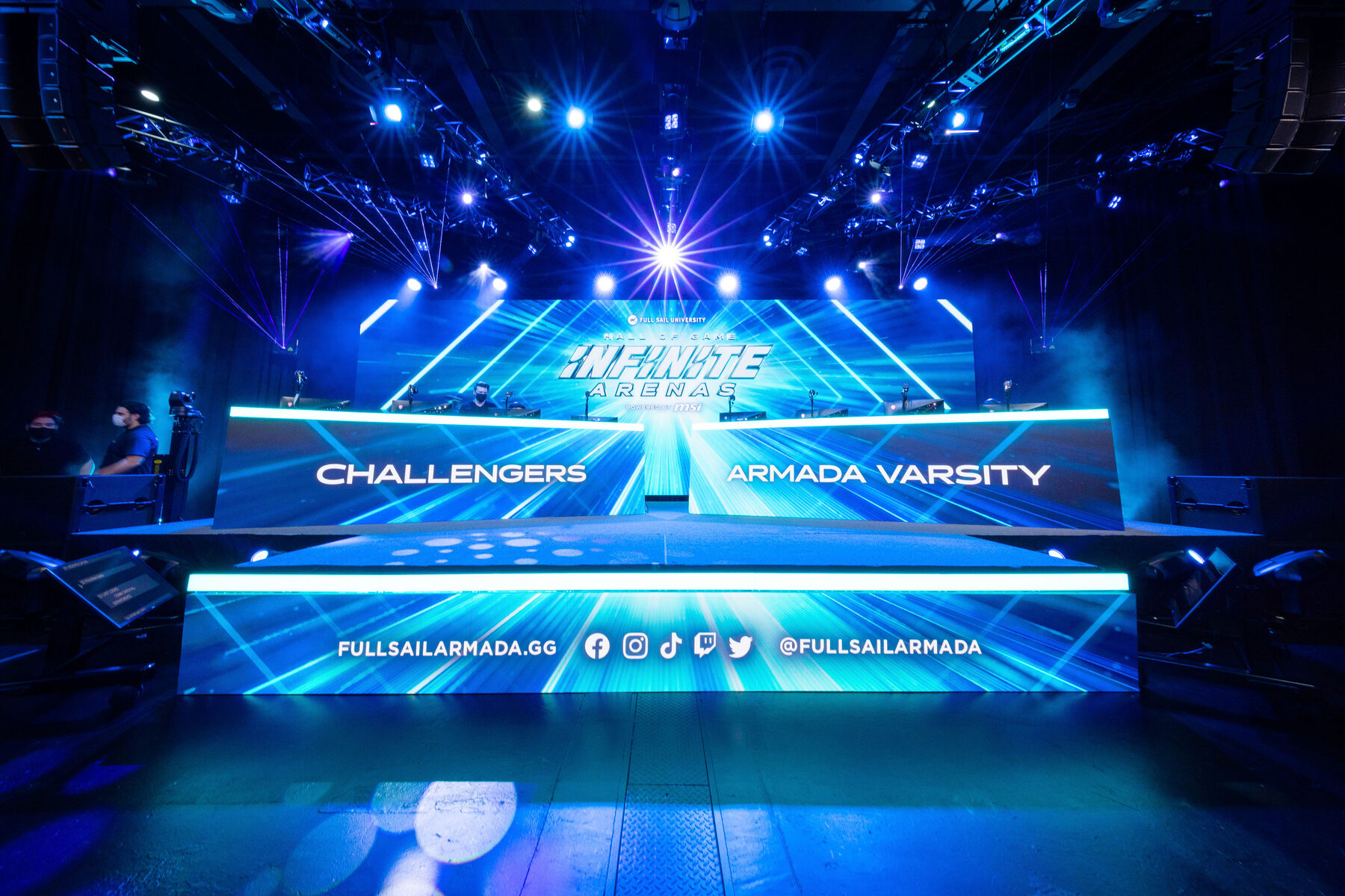 The Infinite Arenas stage head-on: the logo on a wide LED wall above two team desks labeled Challengers and Armada Varsity, lit in blue