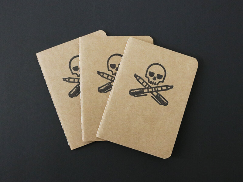Three kraft pocket notebooks stamped with a skull over crossed pencil and marker
